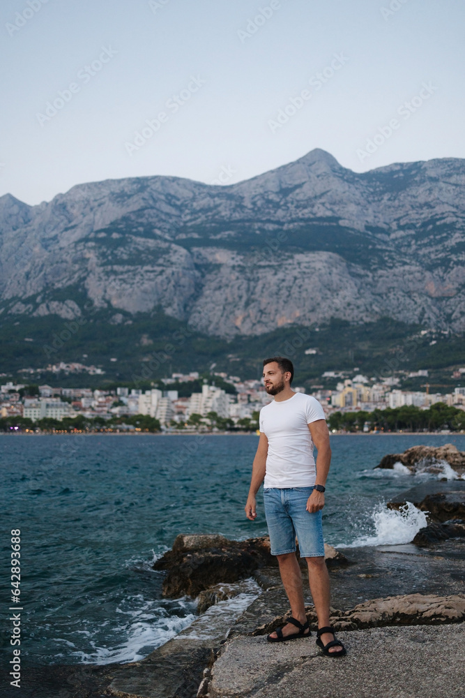 Handsome bearded man stand on the edge of a rock and look at the sea sunset. Back view of big mountains