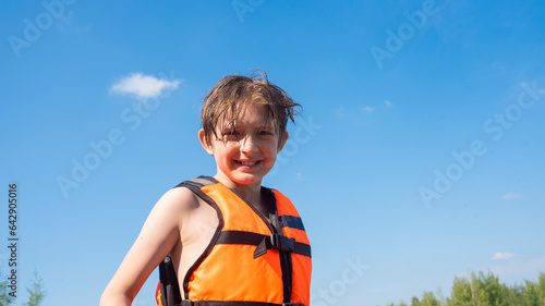 portrait of a happy boy in an orange life jacket in summer. joyful 10 year old smiling child against the blue sky in an orange vest.
