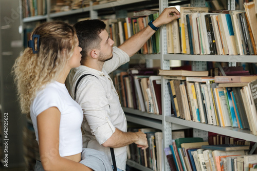 Man take book from book shelf to show his girlfriend. Classmates in college library