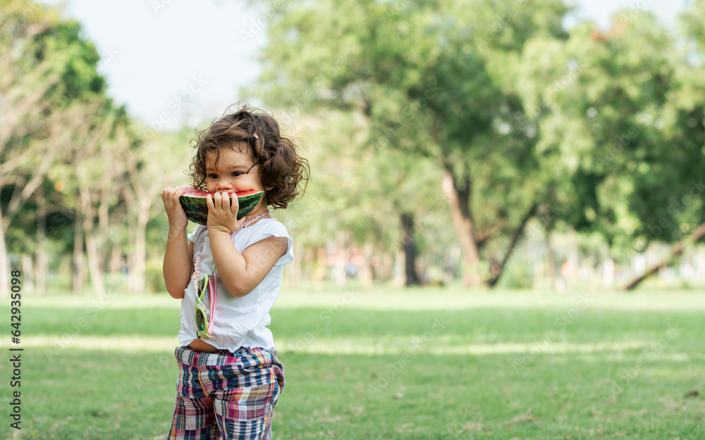 Portrait little caucasian cute girl playing, eating piece of watermelon with happiness while standing on the green field in park. Fruits, Food and Education Concept. Portrait little caucasian cute girl playing, eating piece of watermelon with happiness while standing on the green field in park. Fruits, Food and Education Concept.