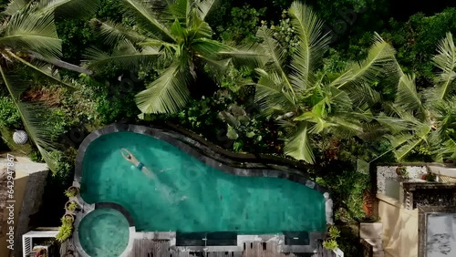 Aerial view of man jumping and dives in infinity pool at jungle and swims under the water. Sporty male swimmer, influencer at luxury villa. Drone