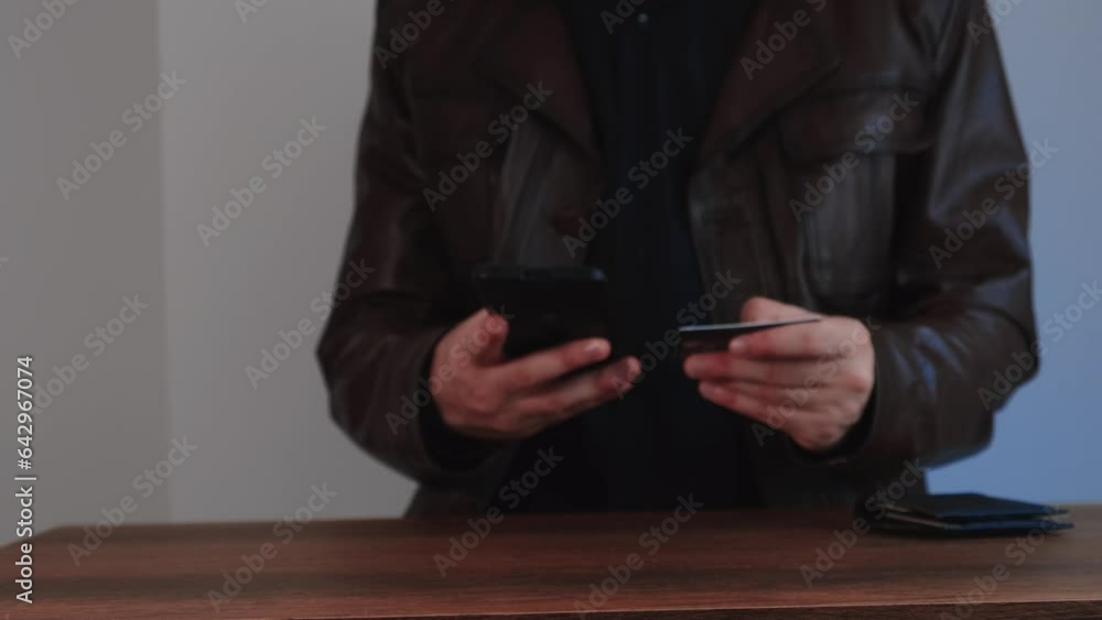 man in leather jacket doing internet shopping at table with phone and credit card in hand man in leather jacket doing internet shopping at table with phone and credit card in hand