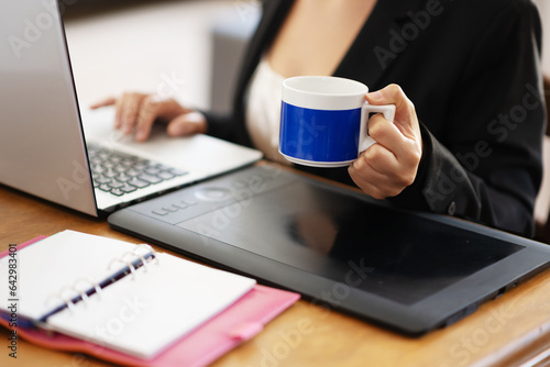 Businesswoman in working environment typing on a laptop and holding a cup of coffee