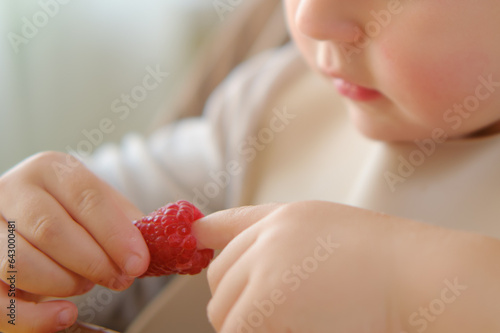 The child is sitting at home and enjoying raspberries for a snack. Kid boy aged two years (two-year-old)