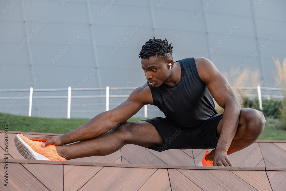 Naklejka premium Confident young African man in sportswear doing stretching exercises while training outdoors