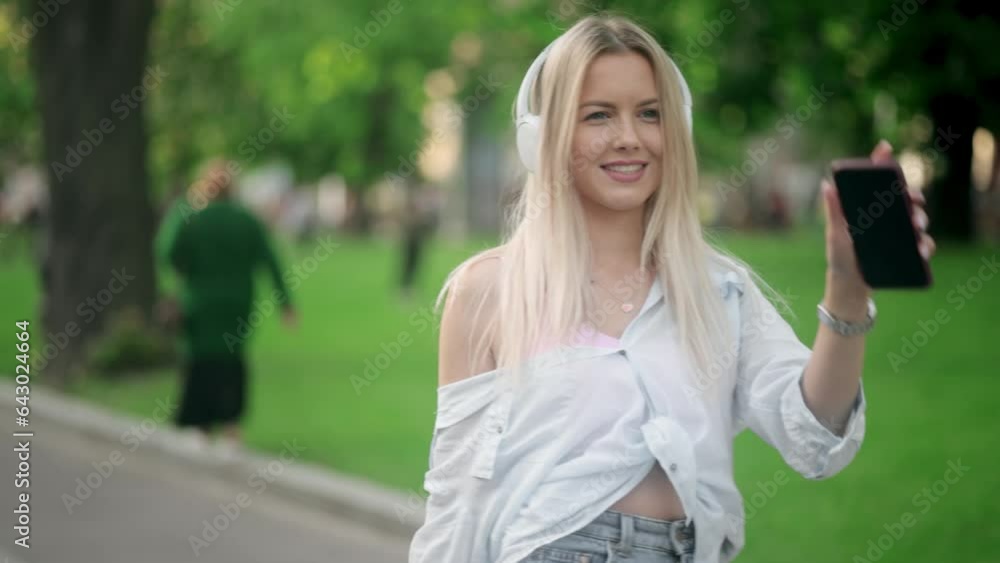 Charming young blond woman walking down the street with headphones listening to music and looking around outdoors Happy relaxed lady walking on the city centre enjoying beautiful day alone Charming young blond woman walking down the street with headphones listening to music and looking around outdoors Happy relaxed lady walking on the city centre enjoying beautiful day alone