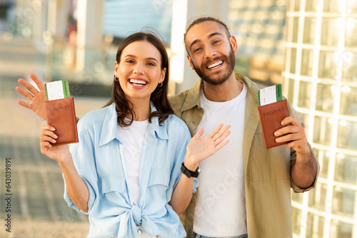 Happy young european guy with beard and woman hold passports and tickets, enjoy trip to station photo