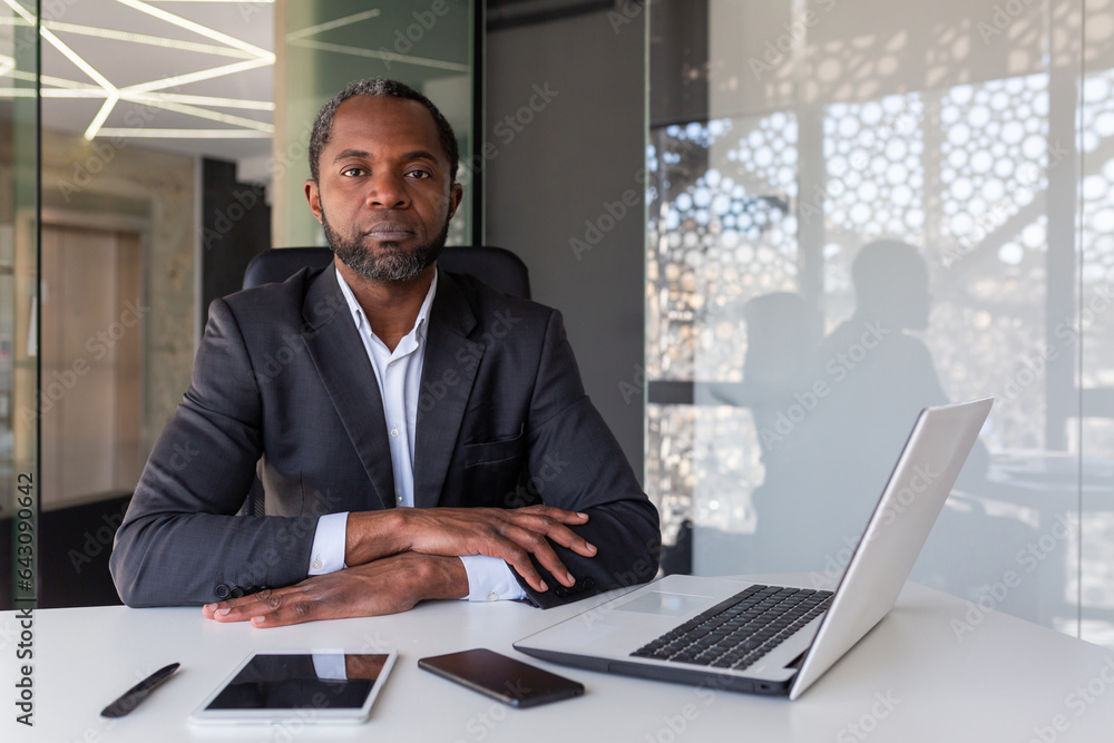 Φωτογραφία Portrait of serious thinking and concentrated boss, african american man looking at camera with folded hands on desk, boss working with laptop inside office, investor confident of victory