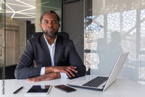 Portrait of serious thinking and concentrated boss, african american man looking at camera with folded hands on desk, boss working with laptop inside office, investor confident of victory.