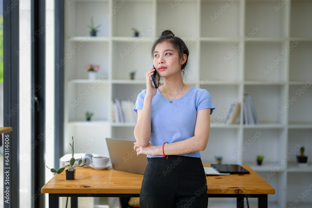 Asian young woman standing using mobile phone to call someone at desk in work area. Looking outside the window. Asian young woman standing using mobile phone to call someone at desk in work area. Looking outside the window.