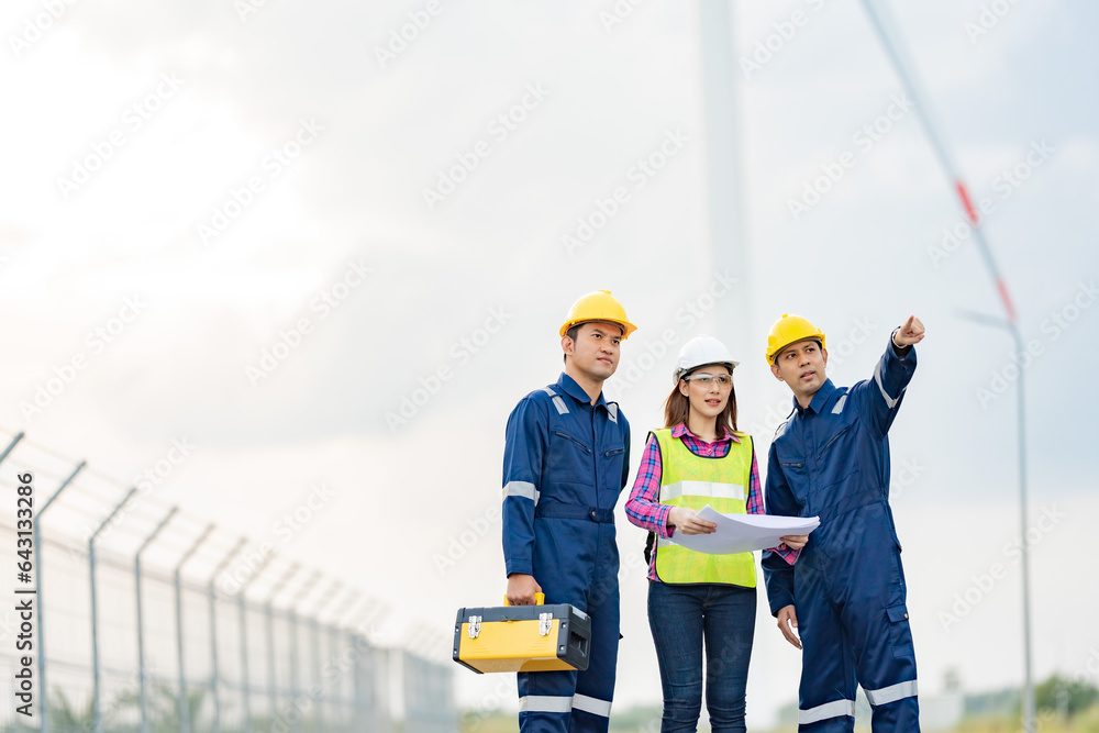A team of 3 male and female engineers wearing vests, hard hats and mechanics with toolboxes. spread out blueprint Looking and pointing ahead There is wind turbine power. Near industrial factories A team of 3 male and female engineers wearing vests, hard hats and mechanics with toolboxes. spread out blueprint Looking and pointing ahead There is wind turbine power. Near industrial factories