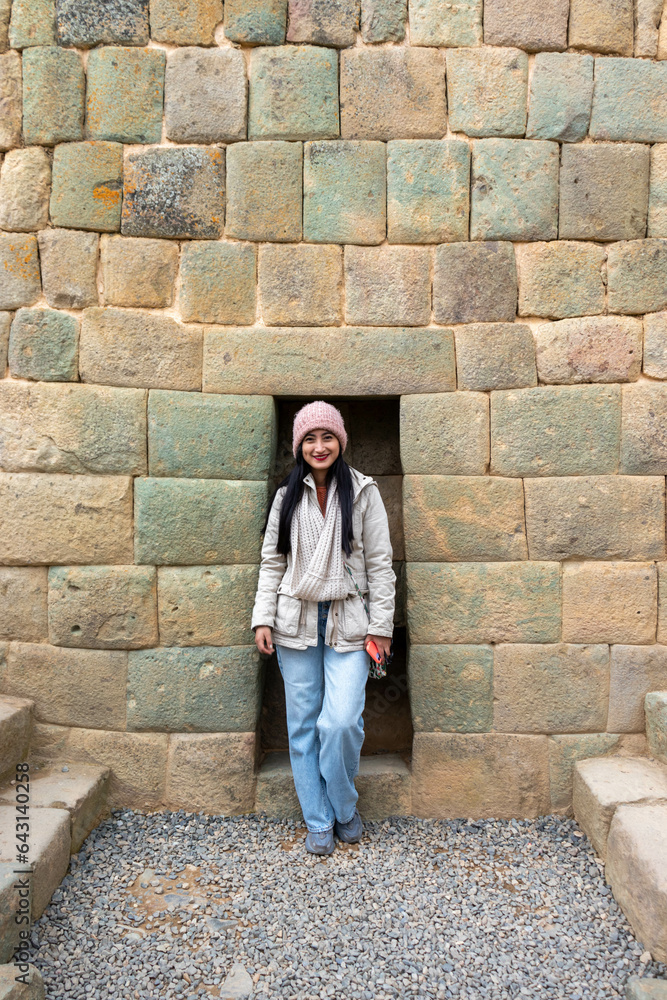 Mujer joven abrigada parada frente a una pared de piedras incas  - obrazy, fototapety, plakaty 
