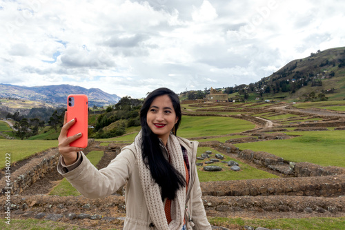 turista joven en las ruinas de ingapirca realizandose una autofoto con su teléfono movil y sonriendo muy alegre en una tarde de primavera en las montañas de los andes  photo