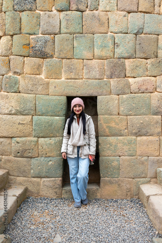 Mujer joven abrigada parada frente a una pared de piedras incas 