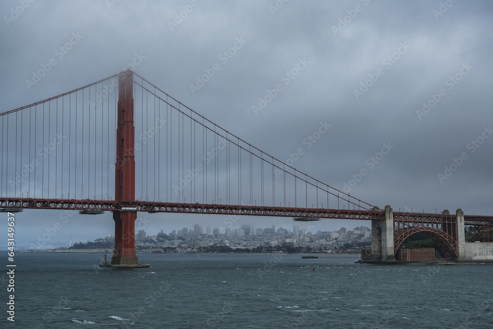 Red metal steel suspension bridge Golden Gate Bridge in San Francisco Bay Area with coast and city downtown skyline silhouette on cloudy day from outdoor cruiseship cruise ship deck