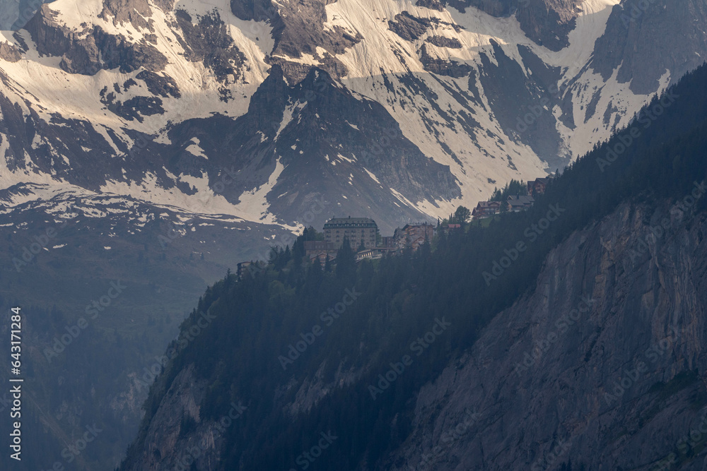 Buildings Sitting Along a Cliff of the Swiss Alps in Switzerland in the Summer with Mountains Peaking Through Clouds in the Background with Houses in the Foreground