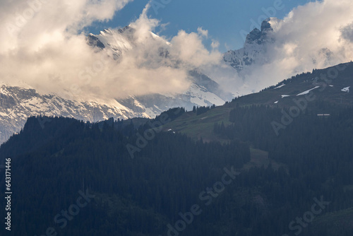 Buildings Sitting Along a Hill of the Swiss Alps in Switzerland in the Summer with Mountains Peaking Through Clouds in the Background