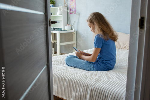 A cute teenage girl is watching videos or writing messages on her smartphone while sitting on the bed in the bedroom. Communication of modern children