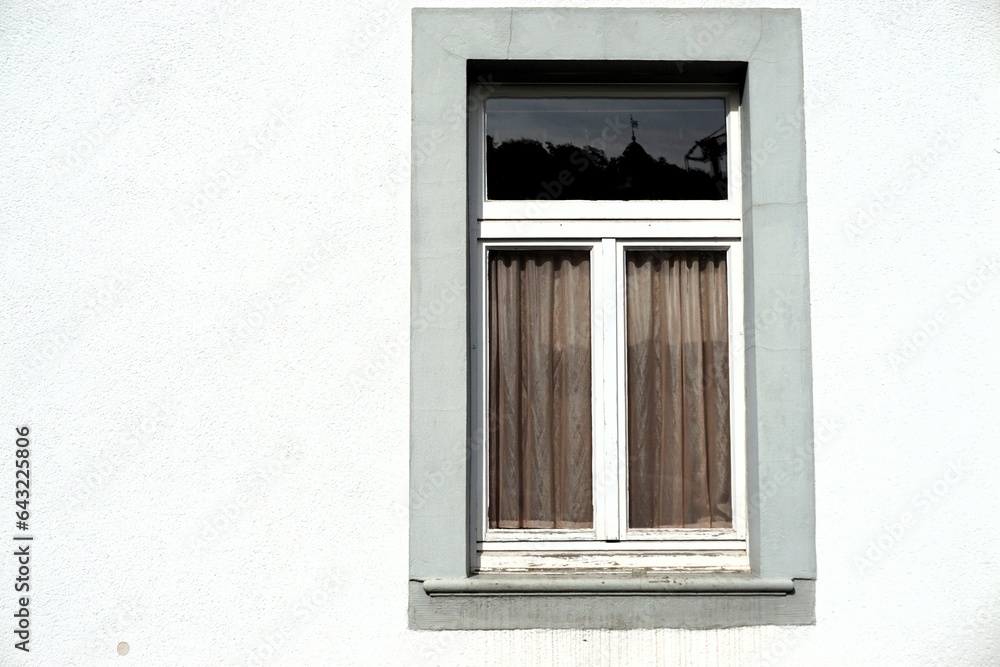 Altes Fenster mit weißem Kreuzrahmen aus Holz und grau-braunen Stoffvorhängen hinter hinter Glasscheibe an weißer Hausfassade bei Sonne am Mittag im Sommer