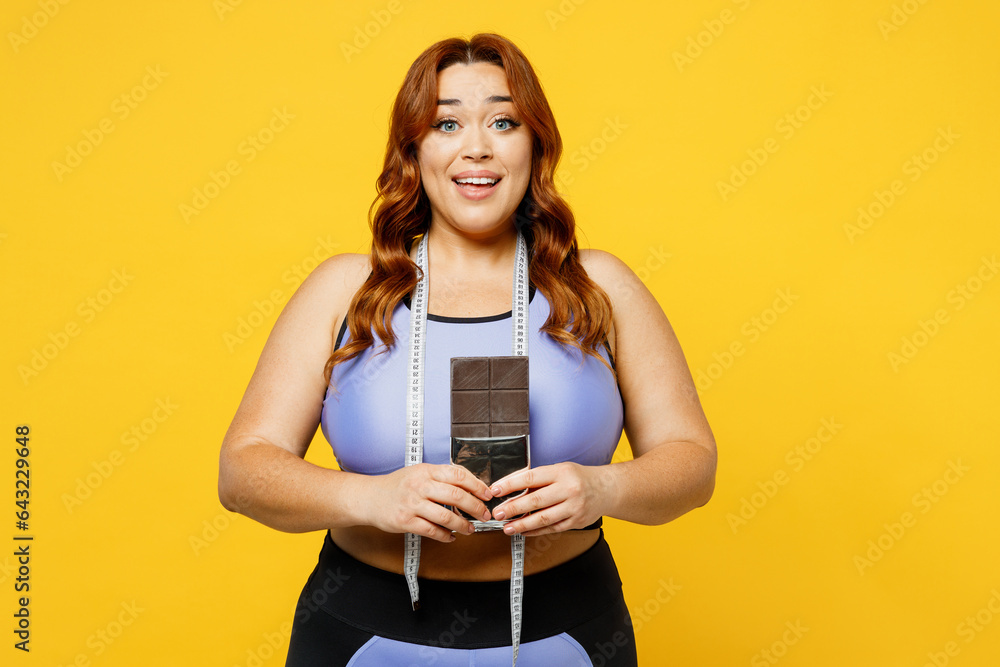 Young happy chubby overweight plus size big fat fit woman wear blue top warm up training hold bar of chocolate measure tape isolated on plain yellow background studio home gym. Workout sport concept. Young happy chubby overweight plus size big fat fit woman wear blue top warm up training hold bar of chocolate measure tape isolated on plain yellow background studio home gym. Workout sport concept.