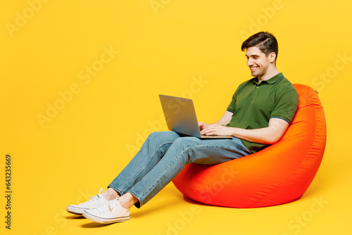 Full body young programmer happy IT man wears green t-shirt casual clothes sit in bag chair hold use work on laptop pc computer isolated on plain yellow background studio portrait. Lifestyle concept.