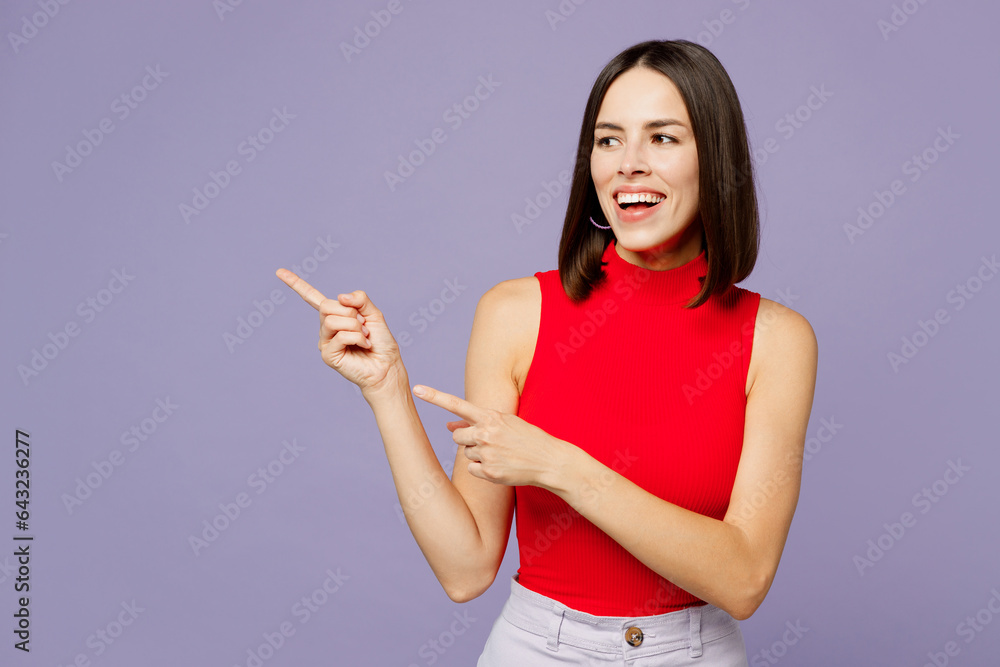 Young smiling woman she wears red tank shirt casual clothes pointing index finger aside indicate on workspace area copy space mock up isolated on plain pastel light purple background studio portrait. Young smiling woman she wears red tank shirt casual clothes pointing index finger aside indicate on workspace area copy space mock up isolated on plain pastel light purple background studio portrait.