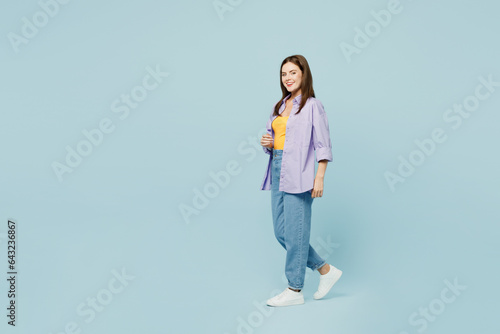 Full body side view young happy woman she wearing purple shirt yellow t-shirt casual clothes walk going look camera isolated on plain pastel light blue background studio portrait. Lifestyle concept.