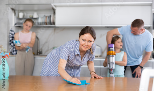 Smiling tidy young housewife cleaning apartment together with her husband and daughters, assiduously wiping kitchen table