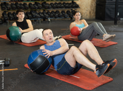 Focused adult man taking part in high-intensity group training session, doing V-sit exercise with medicine ball and twisting torso to strengthen abdominal and oblique muscles.. © JackF