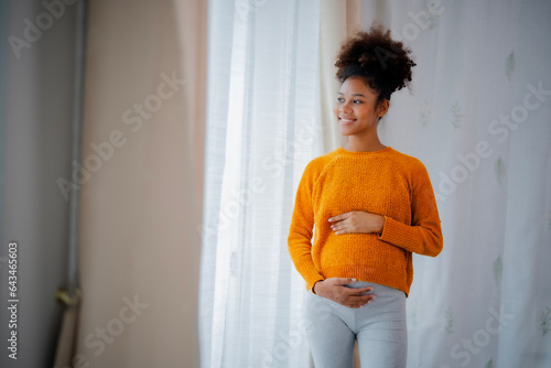 African American pregnancy woman in yellow sweater and gray legging standing beside glasses door, embrassing her belly, looking outside with smile of happiness. photo