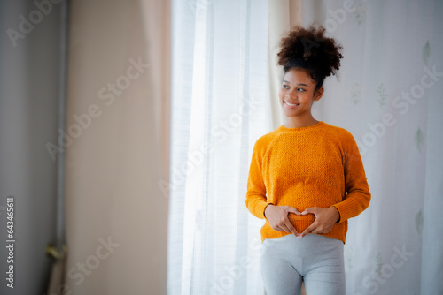 African American pregnacy woman standing beside glasses door, made her hand in heart shape at her belly looking outside window with smile of love and happiness. photo