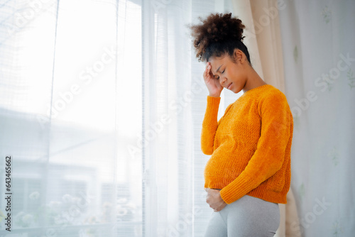 African American pregnancy woman standing beside glass window, hands at head and belly, eyebow fold together, being stress or had headache with white curtain copy space. photo