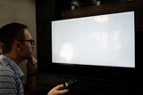 Man watching television, turning on plasma flatscreen TV-set, pointing remote control at empty TV screen. Guy switching channels at home. Mockup