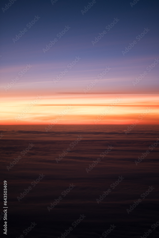 A radiant sunset, as seen from an airplane high above the clouds. The sky is a gradient of bluish purple and reddish orange. The light reflects off the tops of the clouds.