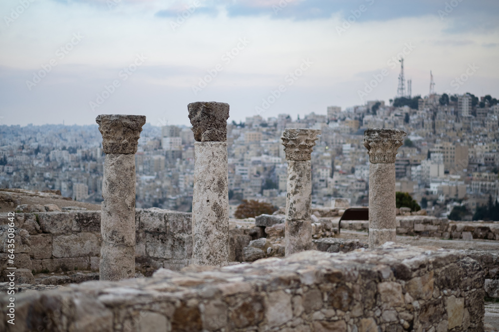 In the background, the modern cityscape of Amman covers rolling hills. In the foreground, ancient classical columns, the ruins of a Roman temple, are weathered but stand tall atop the Amman Citadel.