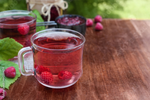 Hot raspberry tea in two transparent cups on a wooden table with blurred green background. Fresh berries, leaves and jars of jam. Side view, place for text copy space.