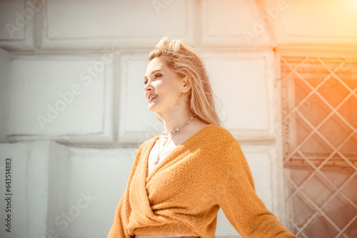 A middle-aged woman looks like a good blonde with curly beautiful hair and makeup on the background of the building. She is wearing a yellow sweater.