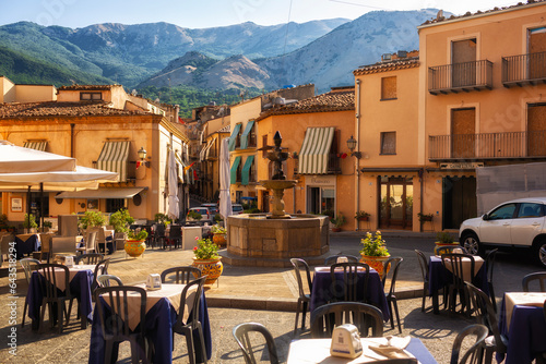 Fototapeta Naklejka Na Ścianę i Meble -  Castelbuono, ITALY - August, 4, 2023. Fountain and cafes with tables in the small town square - piazza of Castelbuono, Sicily. Palermo Province, Madonie mountains on background.