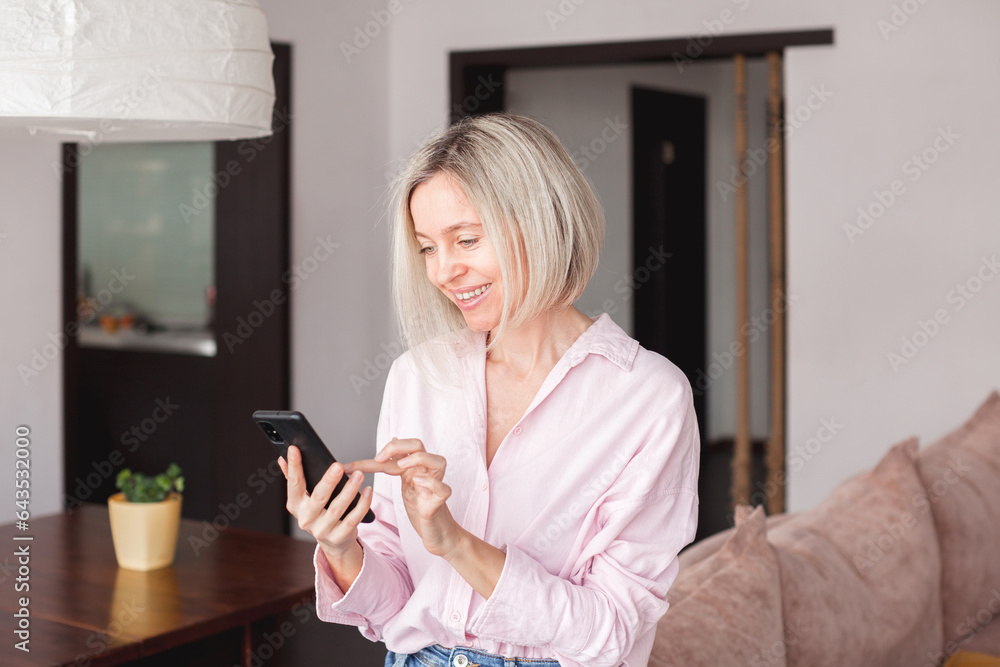 Attractive middle-aged 40s woman looking at her smart phone and smiling while standing at home Attractive middle-aged 40s woman looking at her smart phone and smiling while standing at home
