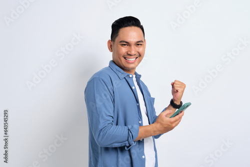 Yes, Great news. Excited young Asian man in casual shirt using smartphone, making winning gesture, celebrating success isolated on white background