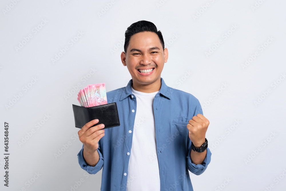 Excited young Asian man in casual shirt holding wallet full of cash, celebrating financial success isolated on white background. Profit and wealth concept Excited young Asian man in casual shirt holding wallet full of cash, celebrating financial success isolated on white background. Profit and wealth concept