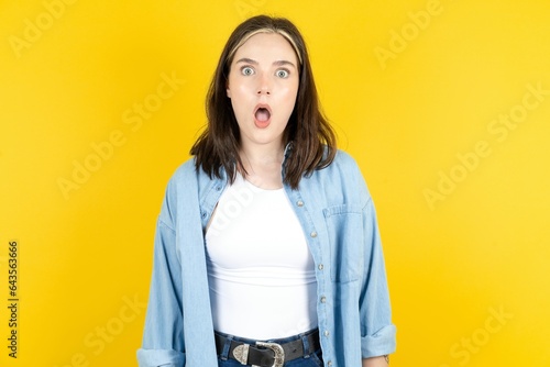Oh my God. Surprised Young beautiful woman wearing denim overshirt stares at camera with shocked expression exclaims with unexpectedness, photo