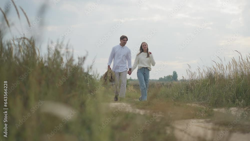 Wide shot young man strolling with woman covering spouse with jacket in slow motion leaving with partner. Happy loving confident Caucasian couple walking in autumn field outdoors Wide shot young man strolling with woman covering spouse with jacket in slow motion leaving with partner. Happy loving confident Caucasian couple walking in autumn field outdoors