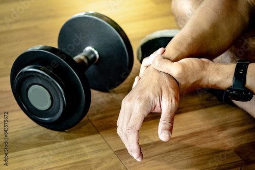 Male athlete massaging his wrist and hand suffering from wrist pain caused by sprain or joint fracture from overweight dumbbell lifting in fitness gym. Sport injury concept photo