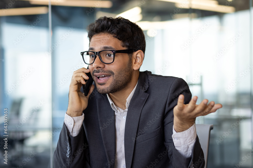 Dissatisfied and upset man talking on phone close up, businessman young in business clothes at workplace inside office. Dissatisfied and upset man talking on phone close up, businessman young in business clothes at workplace inside office.