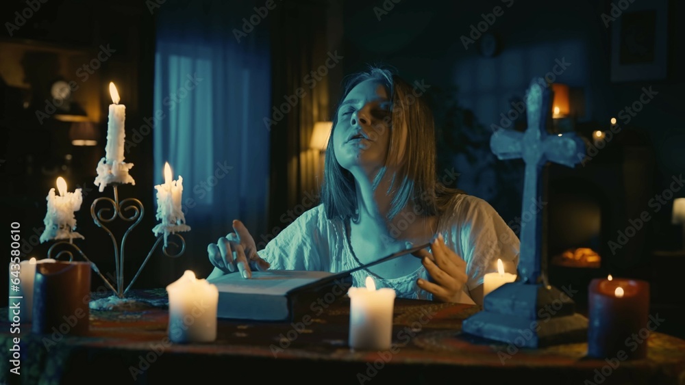 Naklejka premium Shot capturing a young woman sitting at the table, surrounded by candles, crosses and beads, holding an opened witchcraft book, performing a rite, ritual, reading enchantments.