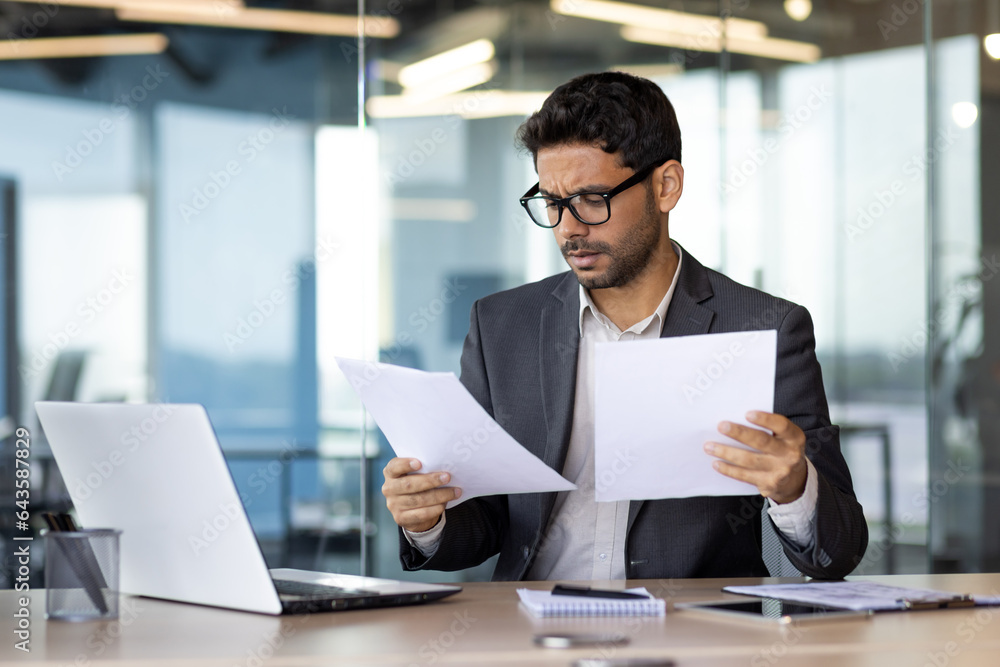 Serious concentrated businessman inside office behind paper work, financier reading and checking reports holding in hands, man thinking about technical solutions at workplace. Serious concentrated businessman inside office behind paper work, financier reading and checking reports holding in hands, man thinking about technical solutions at workplace.