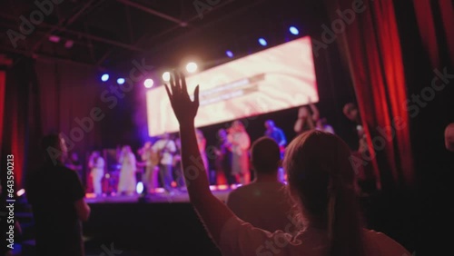 Young girl lifting hand up during christian worship night performing, christian band on the stage singing and playing musical instruments, worshipping God Lord with a crowd, raising hands at concert 