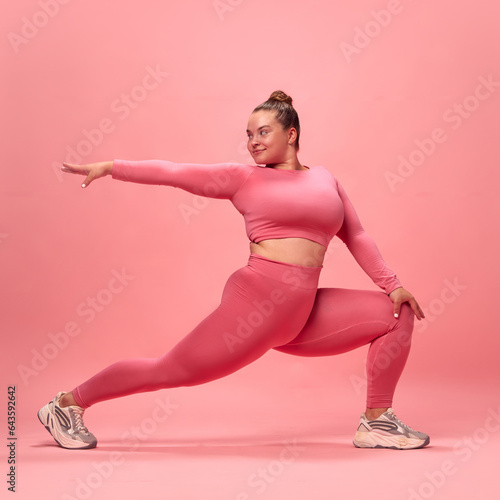 Full body portrait of young plus size sporty woman in pink top and legging training, stretching legs and hands isolated studio background.