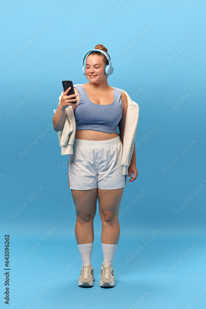 Full length portrait of young smiling plus-size woman staying in sportswear with phone in headphones against blue studio background. Music for workout Full length portrait of young smiling plus-size woman staying in sportswear with phone in headphones against blue studio background. Music for workout